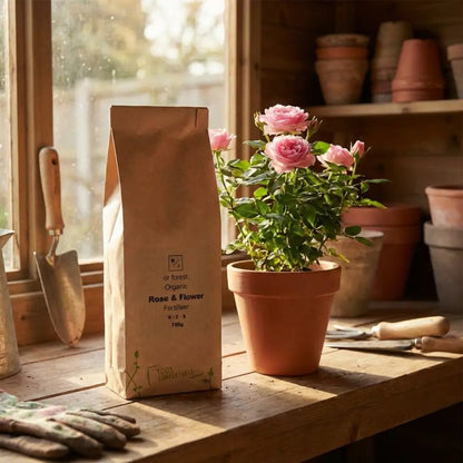 Organic Rose Fertiliser — kraft bag beside potted pink rose in rustic shed window light with tools